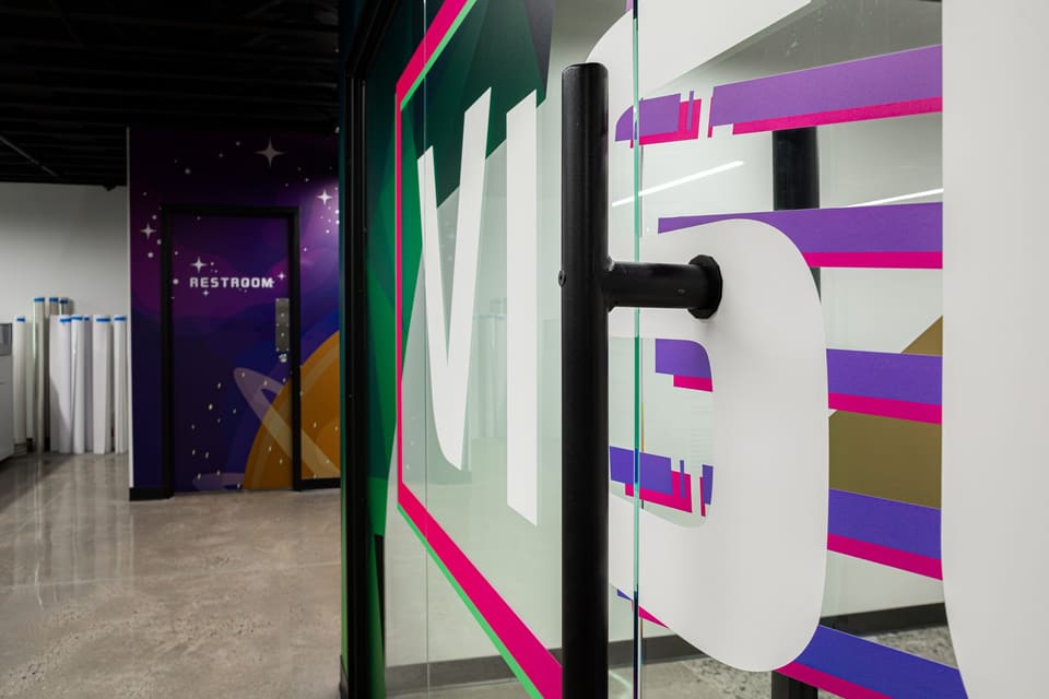 Close view of oversized white lettering and colored stripes cut into a glass wall beside a black door pull