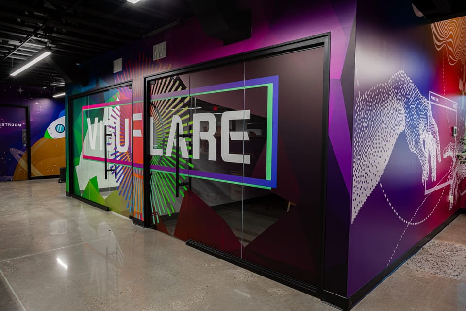 Studio hallway with glass offices carrying lettering and a colorful geometric wall print that wraps around the corner