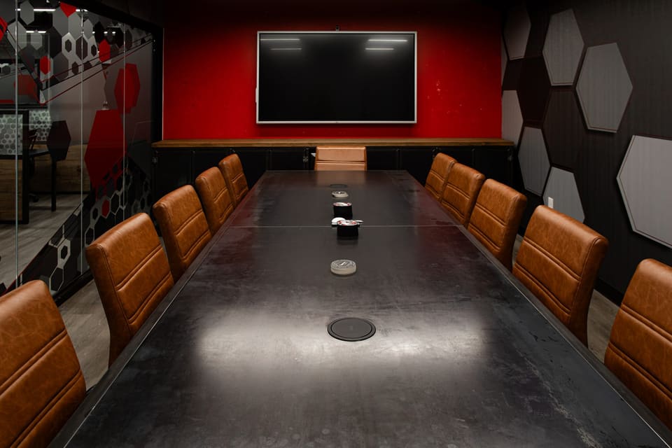 Conference room with a long black table, tan leather chairs, a red wall behind a wall display and gray hexagon tiles