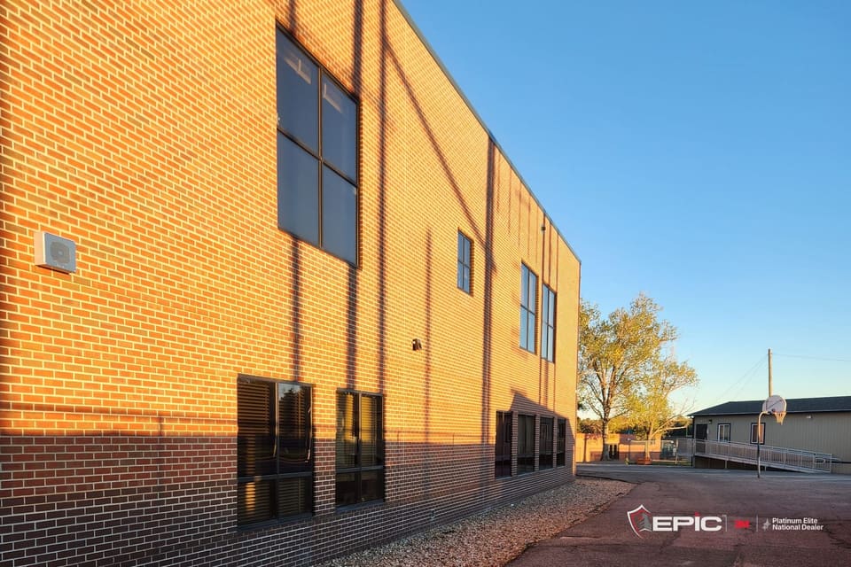 Brick school wall at sunrise, the lower windows dark with exterior solar film, a basketball hoop beyond