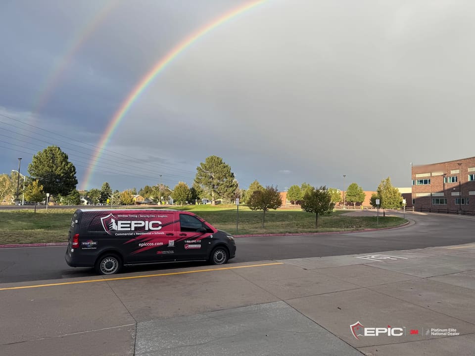 An Epic van parked at the curb of a school drive under a double rainbow, trees and a brick school building beyond