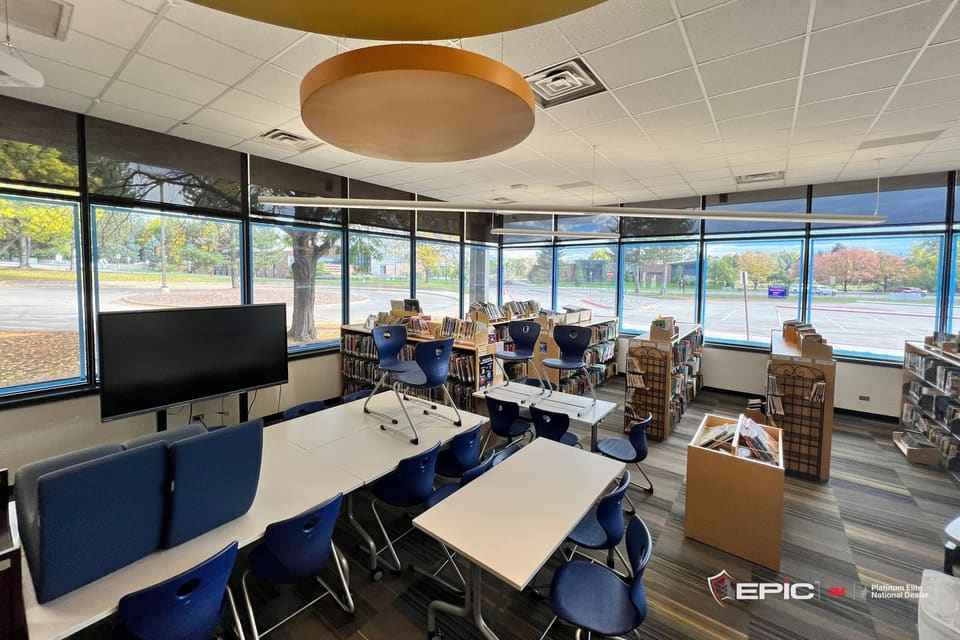 A school library with its window band taped blue on two walls, chairs up on the tables, book bins on the floor