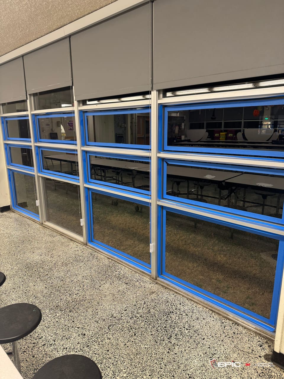A cafeteria window wall at a Dallas ISD school taped in blue, four bays of three lites, lunch tables beyond the glass