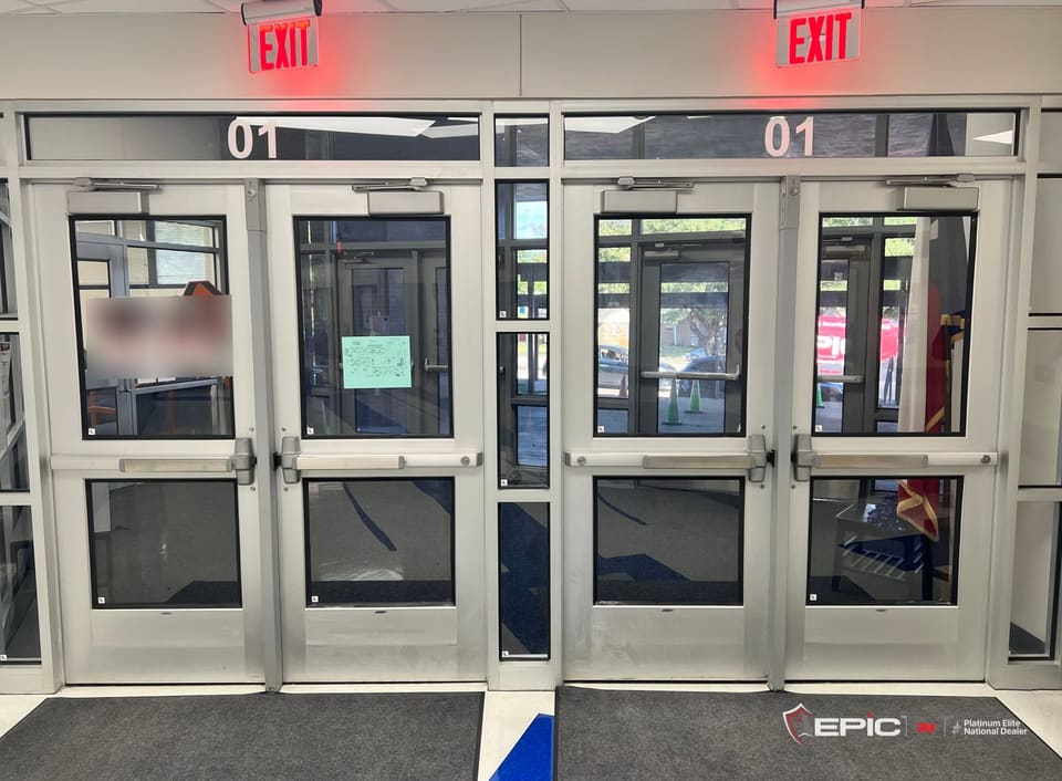 Two pairs of aluminum entry doors at a school, every lite filmed with 3M Ultra S800, the Epic van reflected in the right-hand glass and the transoms marked 01